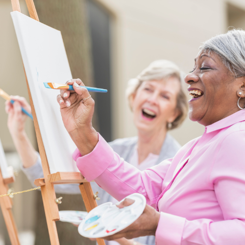 two senior woman laughing together while painting on easel