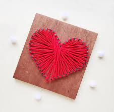 square piece of wood with nails in place to make a heart shape and red string filling in the heart.