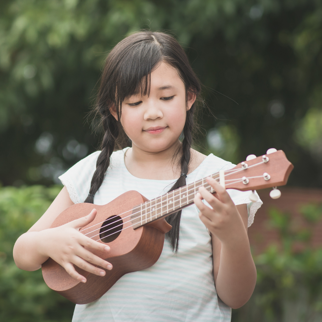 Child with braids playing ukulele with green trees and plants in the background