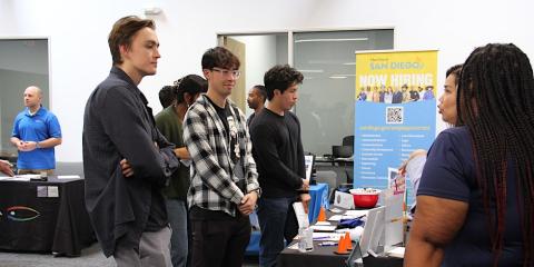 three males standing at a table speaking with woman also standing