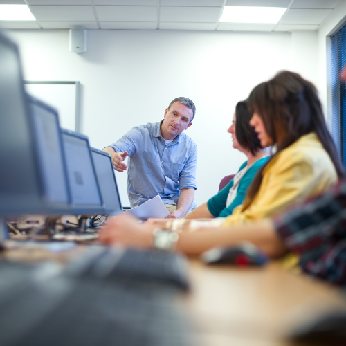 row of computers with students, teach at end of line pointing at computer